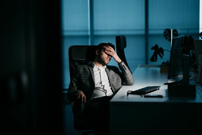 暗いオフィスで疲れ切った男性が目元を手で覆っている