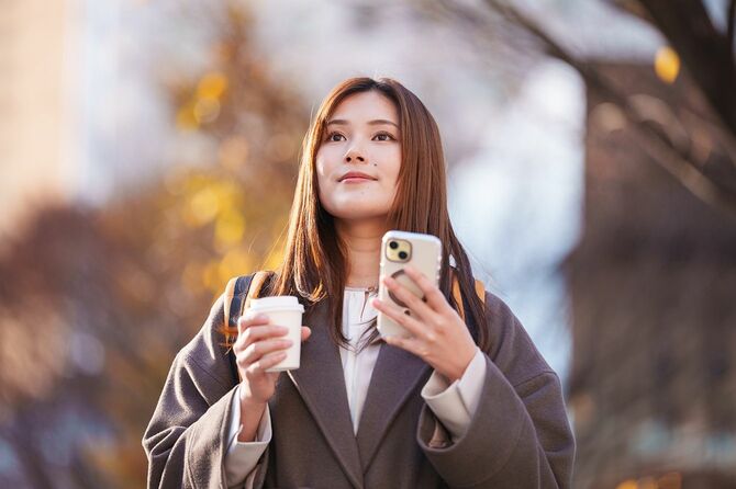 街の屋外でスマートフォンとテイクアウトのコーヒーを手に持つ若い女性