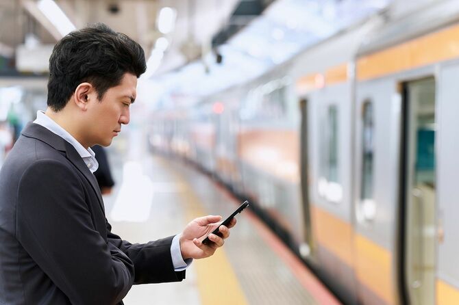 ビジネスマンが駅のホームで電車を待ちながらスマートフォンを見つめている