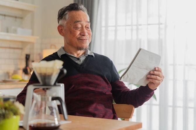 幸せな高齢のアジア人男性がノートパソコンを使い、キッチンでコーヒーカップを持つ