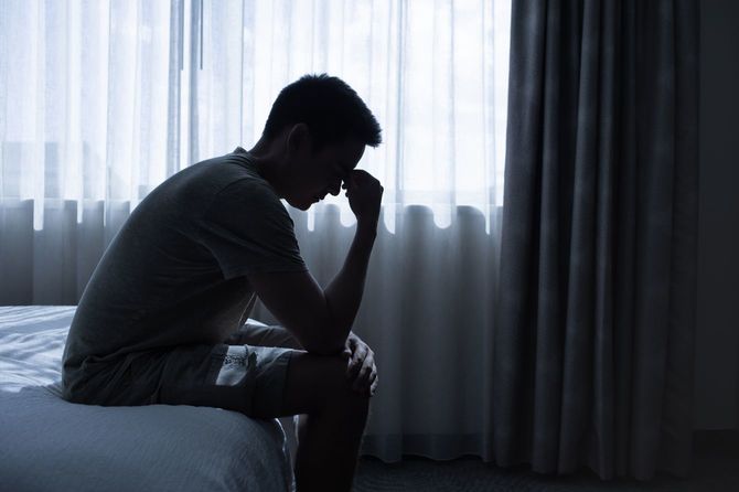 ベッドに腰掛け、眉頭をもんでいる男性のシルエット