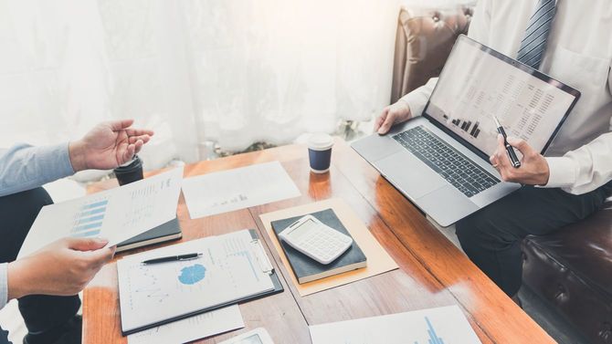 ノートパソコンを開いて数値の説明をする男性に紙資料を手にした男性が質問している