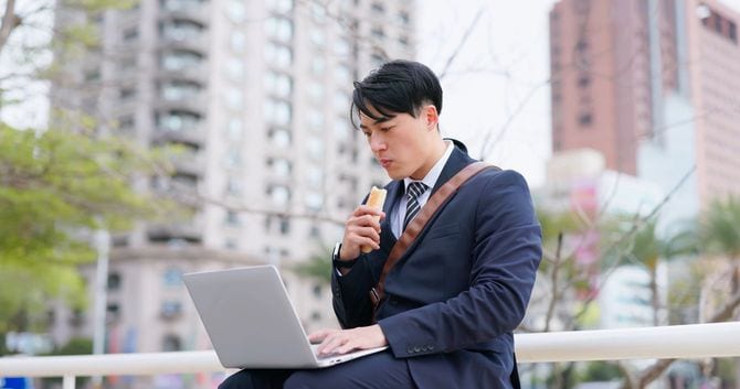 ワンハンドランチを頬張りながらノートパソコンを使用している男性