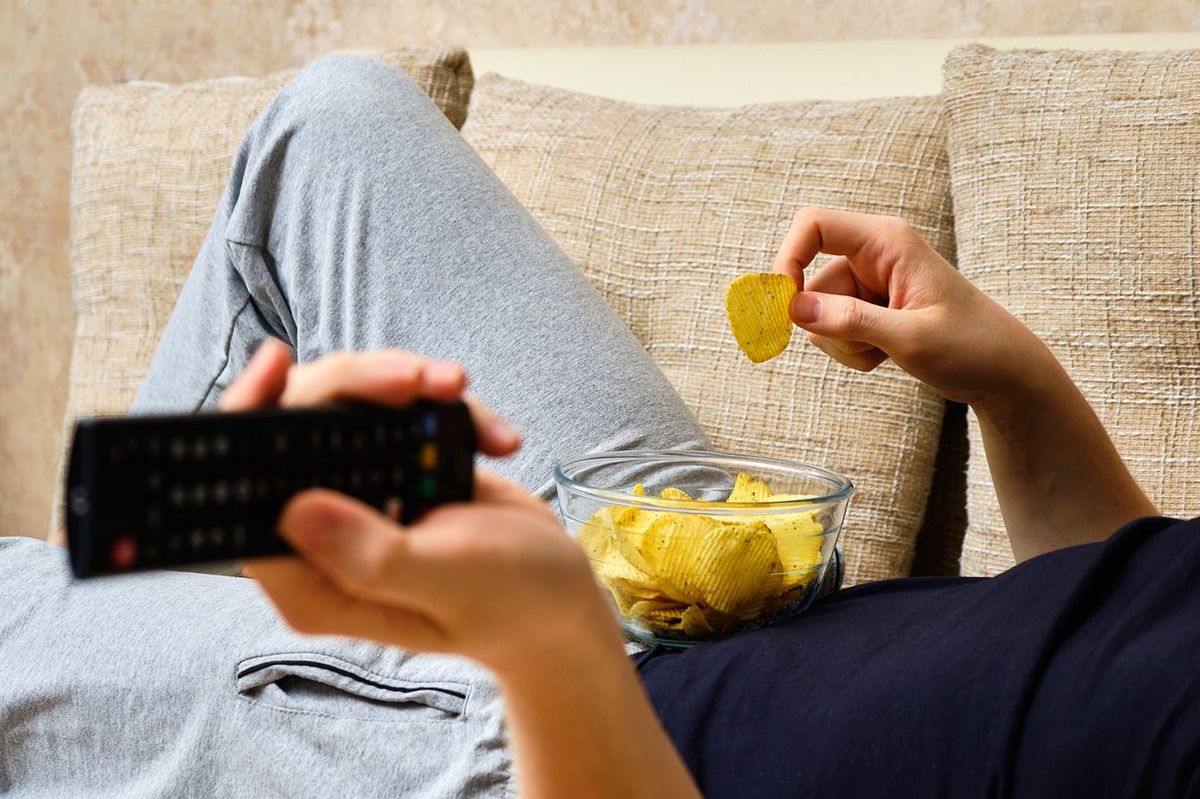 ソファに横たわりポテトチップスを食べながらテレビのリモコンを操作する人