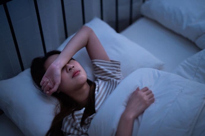 ベッドに仰向けになり、右腕を目に当てて眠ろうとしている女性
