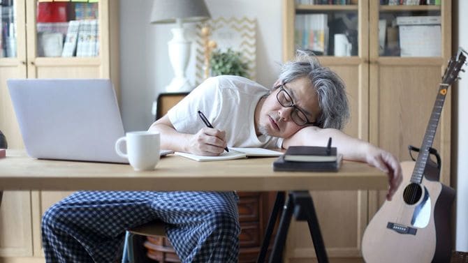 自宅での作業中、疲れて半分机にうつぶせになり目を閉じている男性