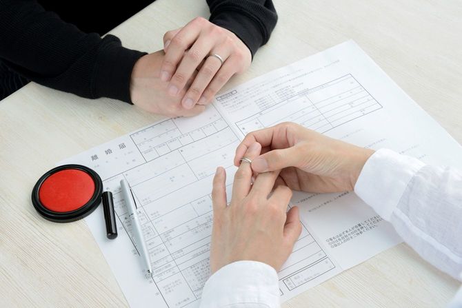 離婚届を挟んで相対して座るカップルの1人が、結婚指輪を外している