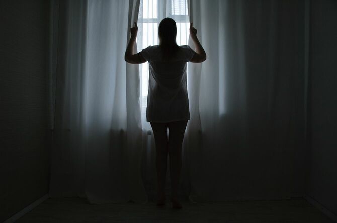 窓辺に立ち朝日が差し込むカーテンを開ける女性