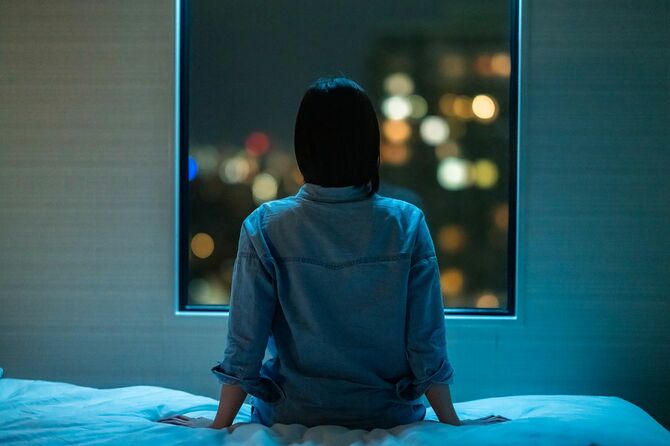 部屋のベッドに一人で座り、夜に窓から覗く女性