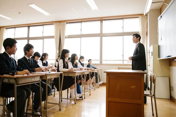 教室で前に建つ先生と話を聞く生徒