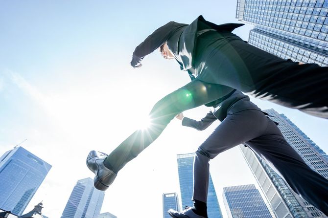 建物に囲まれた青い空の下でジャンプするビジネスパーソンの男