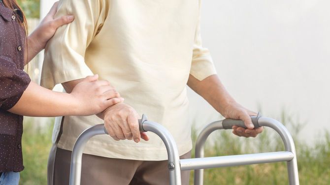 老婦人が手押し車を使用し、介助されながら歩行