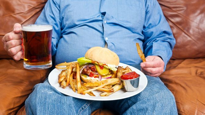 あまりに不健康な食事を膝の上に置き、右手にはビールジョッキを持つ肥満体型の男性