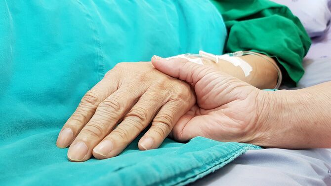 病院のベッドに横たわる高齢者の手を握る人