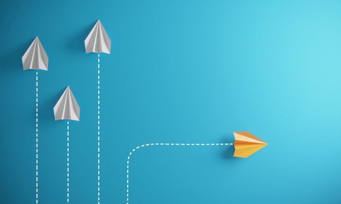 一方向に飛ぶ白い紙飛行機のグループと異なる方向へ飛ぶオレンジ色の紙飛行機