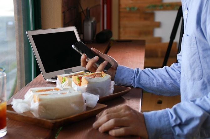 PCとサンドイッチがのったカウンター席でスマホを見ている人