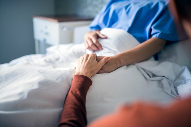 病院のベッドに横になる患者に付き添って手を握っている女性