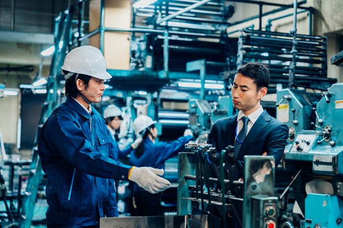 工場で説明する男性の言葉に渋い顔をするスーツ姿の男性