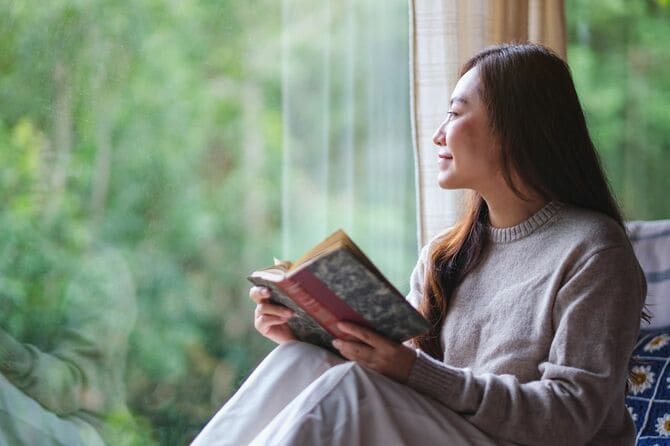 窓辺で本を広げ窓の外を眺める女性