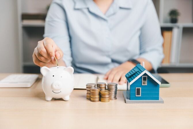 貯金箱にコインを入れる人と積み上げられたコイン、ミニチュアの家