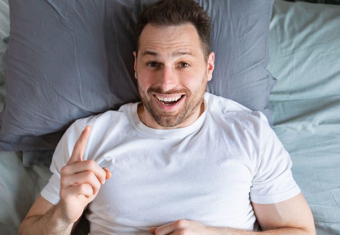 ベッドに横たわって人差し指を立ててひらめくポーズをする男性