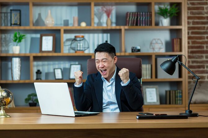 アジアのビジネスマンは喜びで叫び、手を挙げ、勝利を祝い、男性はオフィスの机で働き、ラップトップの画面を見る