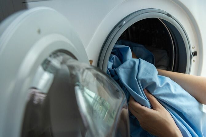 白い洗濯機に汚れた青いシーツを入れる女性の手