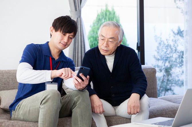 年配の男性にリビングルームでスマートフォンやラップトップの使い方を教えている若い男性