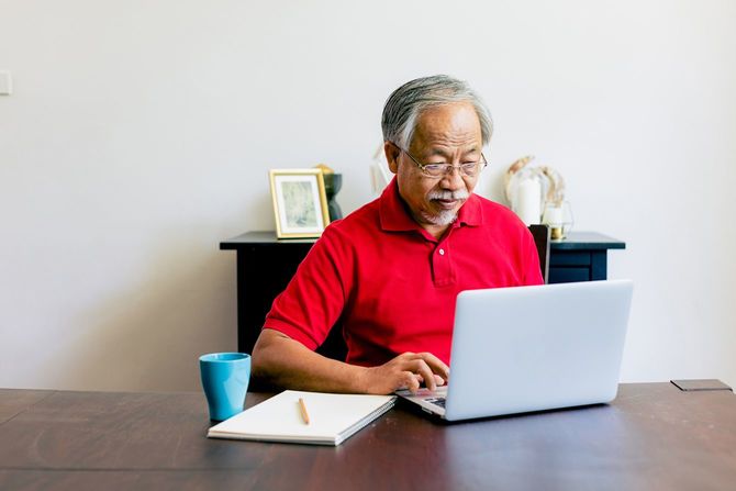 ノートパソコンの横にはノートを置いて、作業するシニア男性