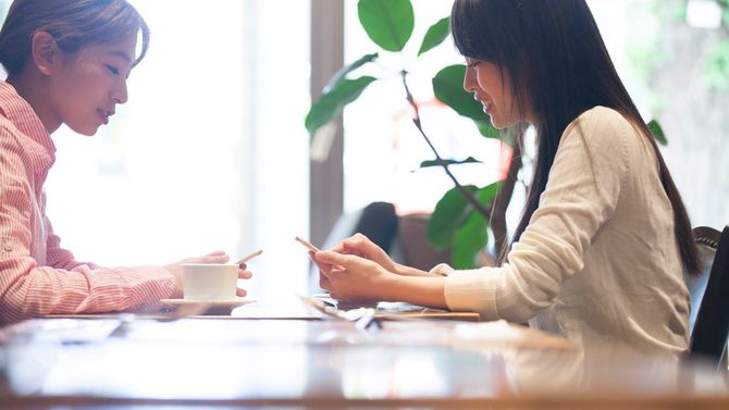カフェで向かい合って座り、スマホを手に会話する女性たち