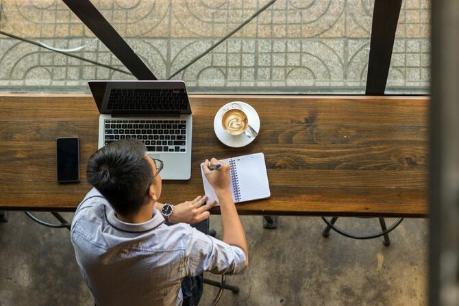 カフェの机でノートパソコンとメモ帳を広げ、メモをしている男性