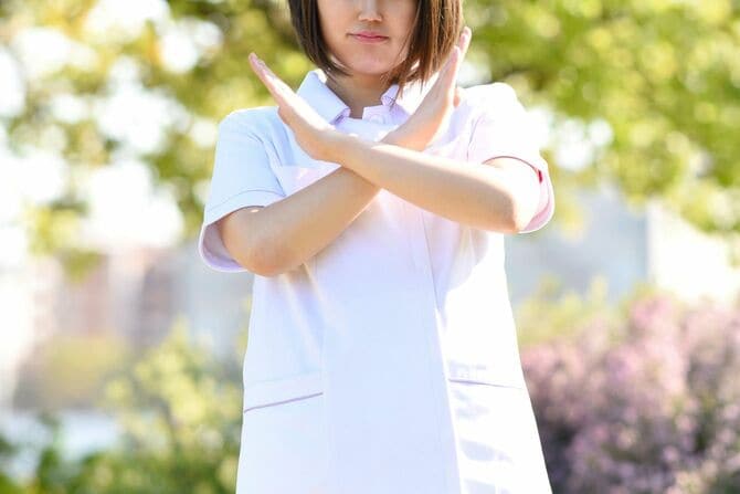 白衣を着て屋外に立ちながら両手で十字のジェスチャーをする医療従事者の女性