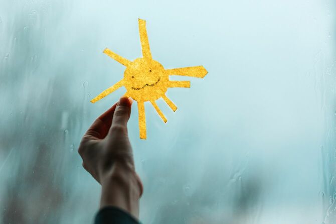 雨が降りつける窓に紙に描いた太陽を付けている手元