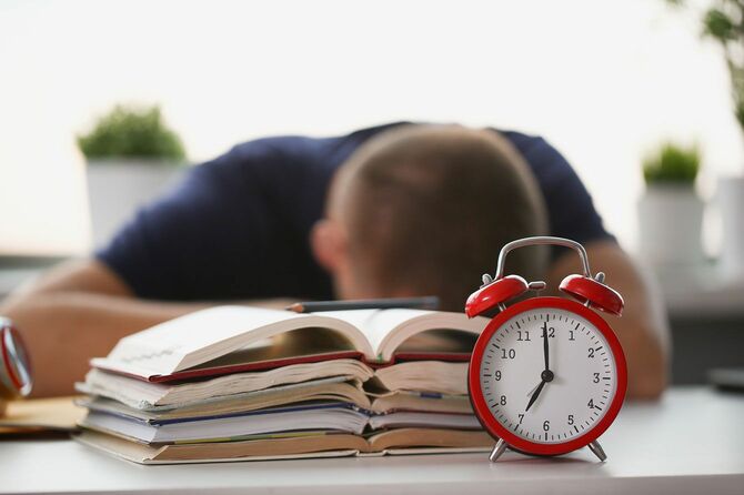 積み上げられた本と目覚まし時計を前に机に突っ伏して寝る男性