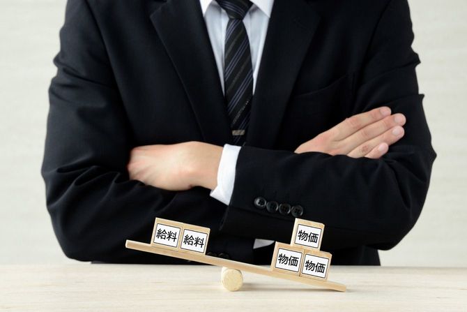 「物価」「給料」と書かれた木のブロックが乗ったシーソーとその後ろで腕を組むビジネスマン