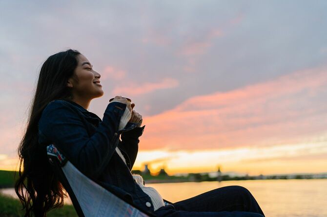 若い女性が湖の近くに座って、日没の間に自然の中で熱い飲み物を飲んで楽しんでいます。