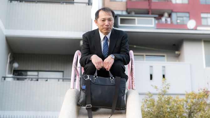 公園の滑り台に座りはにかむビジネスマン