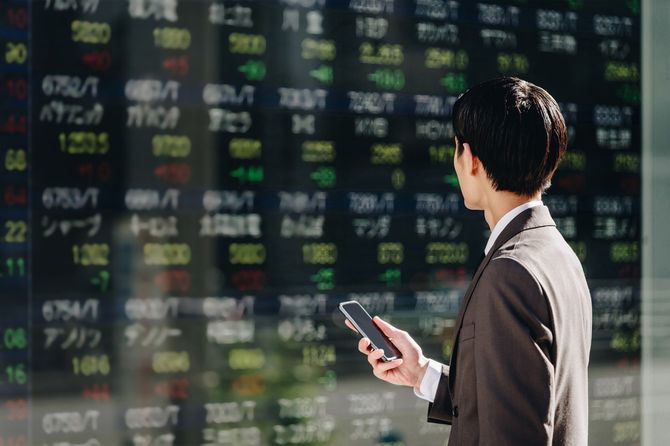 リアルタイム株価ボードの前でスマートフォンで取引する30代の男性