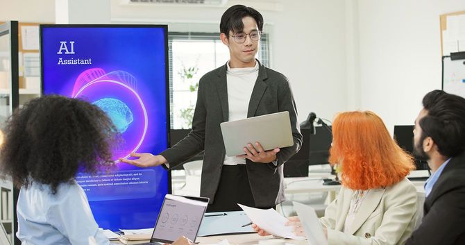 「AIアシスタント」という文字が表示されたディスプレイの前でプレゼンをする男性