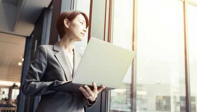 ノートパソコンを手に、オフィスの窓から外を眺めるキャリアウーマン