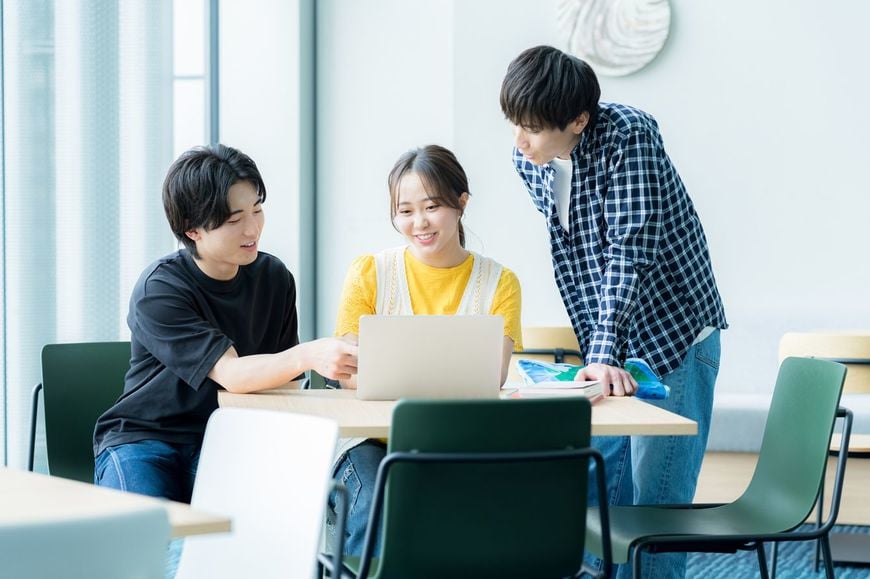 ノートパソコンを見ながら3人の学生が話し合っている