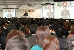 朝到着した順に着席するルール（幹部席除く）。スクリーンには登壇者の顔や資料が映し出される。