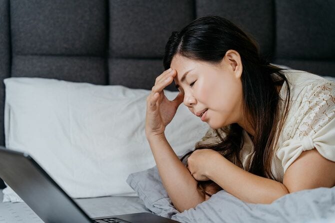 ベッドでノートパソコンを開いて頭を押さえる女性