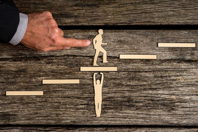 階段を登る男の人形と、階段にぶら下がって落ちそうな男の人形、男性が階段を登る人形を指さしている