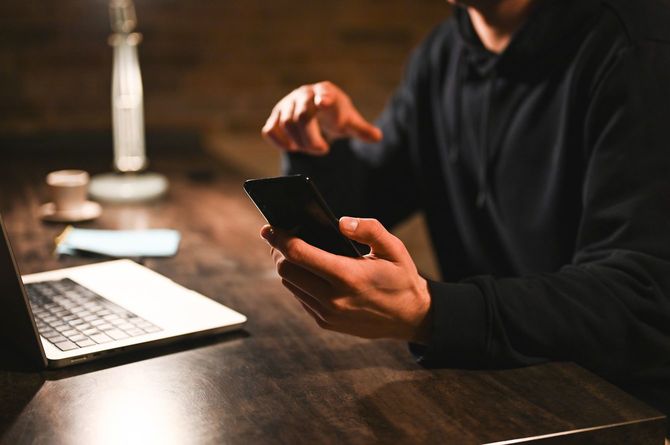 薄暗い部屋でノートパソコンを開きながら、スマートフォンを操作する男性