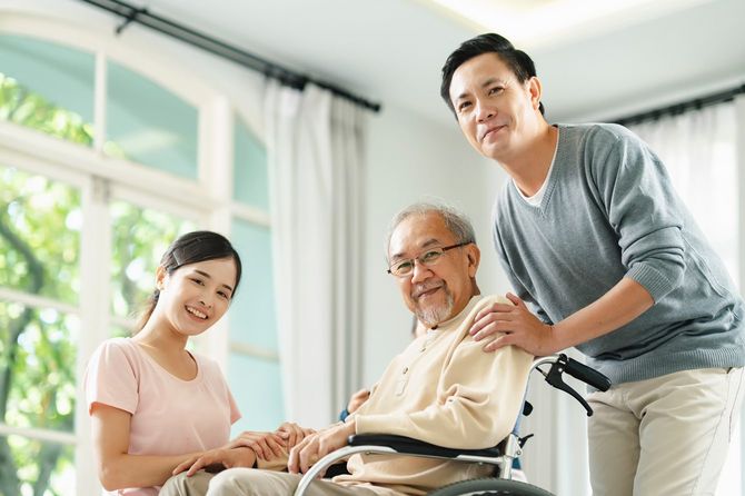 車椅子の高齢男性のそばに立つ男性と座って腕に手を置く女性