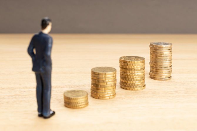 階段状に積み上げられているコインを見つめるミニチュアのサラリーマン