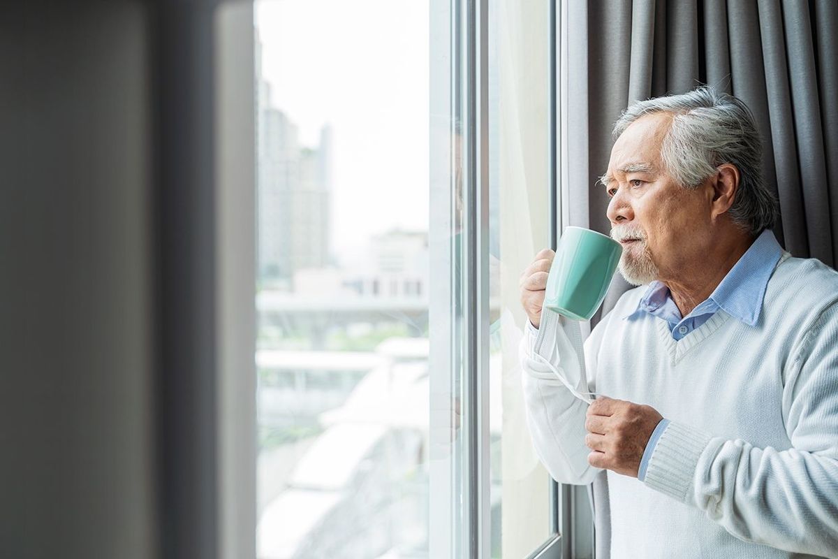 カップを持って窓辺に立ち、外を眺める高齢男性