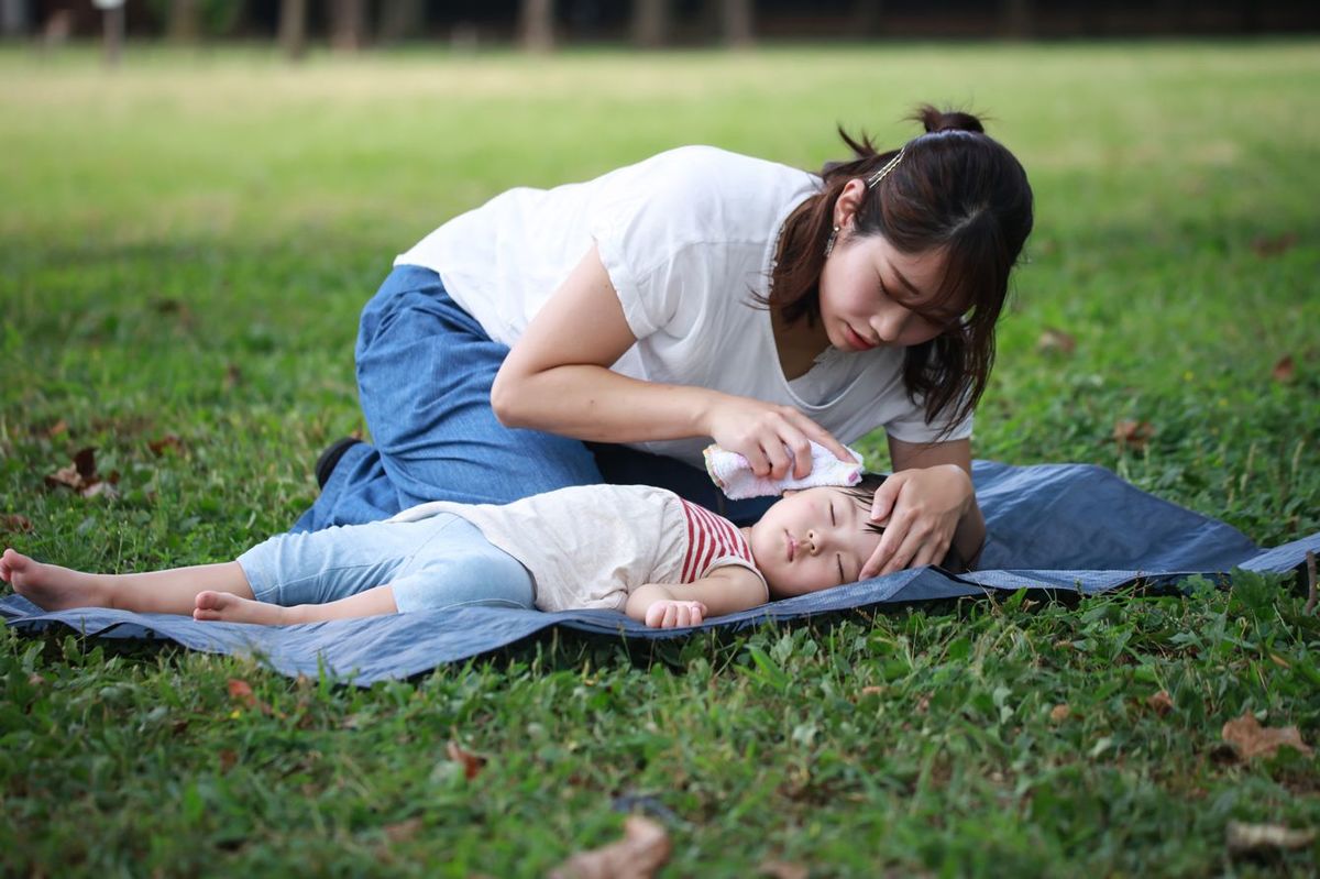 芝生の上に敷いたブルーシートの上で眠る子供の汗をぬぐう母親