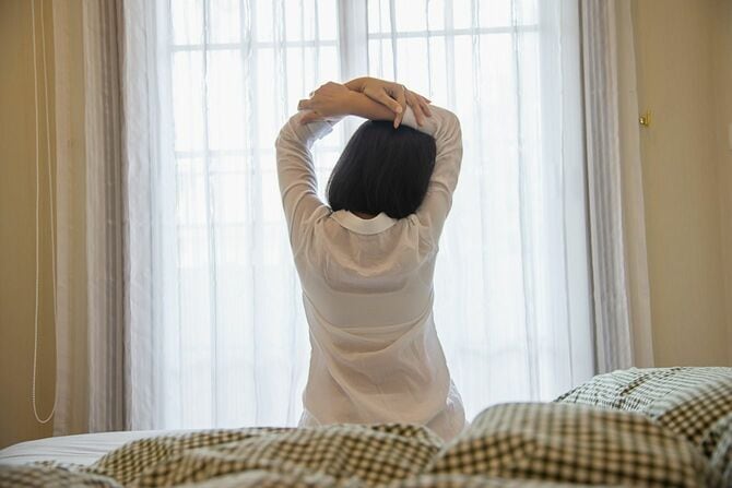 自宅のベッドに座って伸びをする女性の後ろ姿